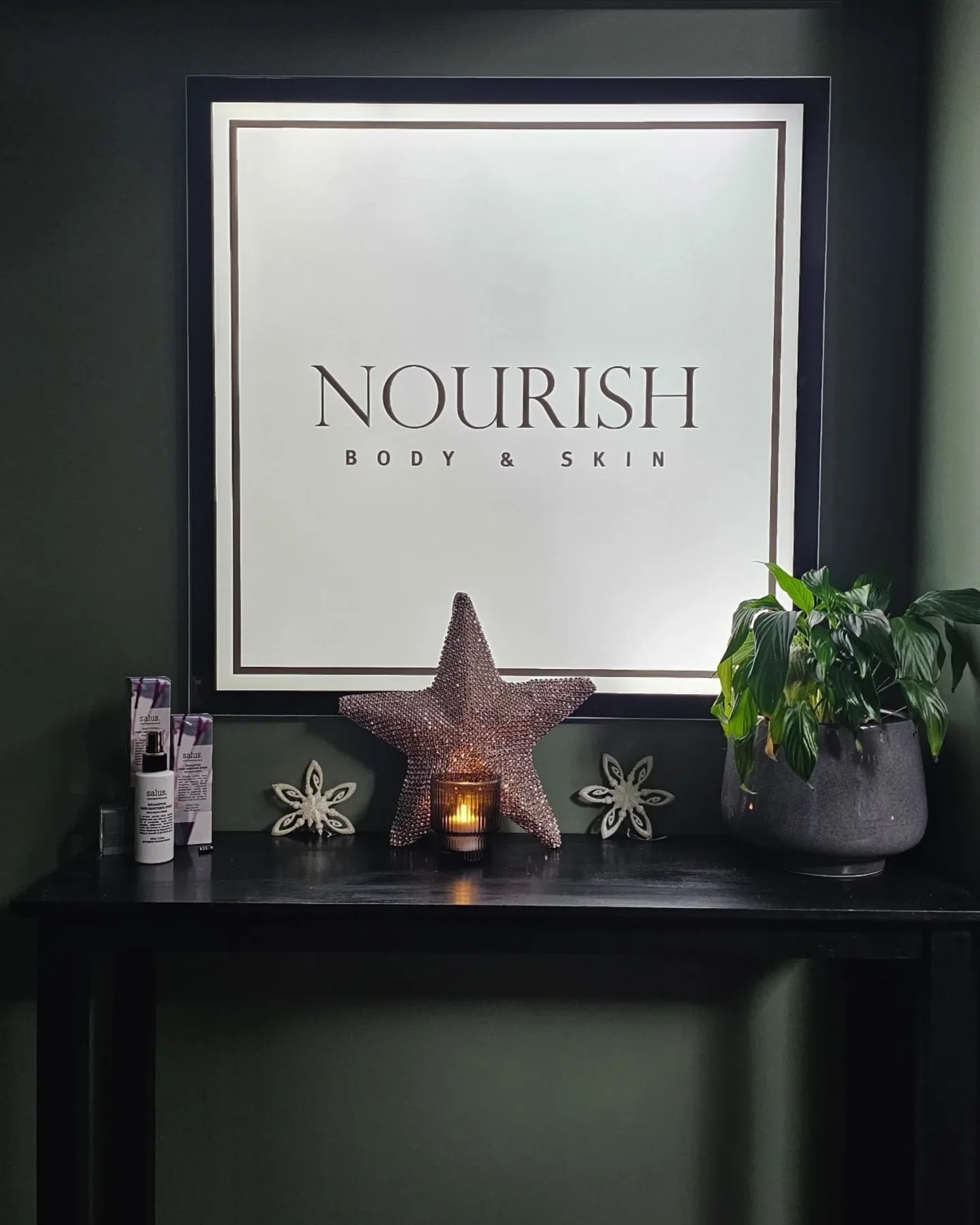 Nourish clinic interior — deep forest-green wall with illuminated brand mirror, candle and peace lily